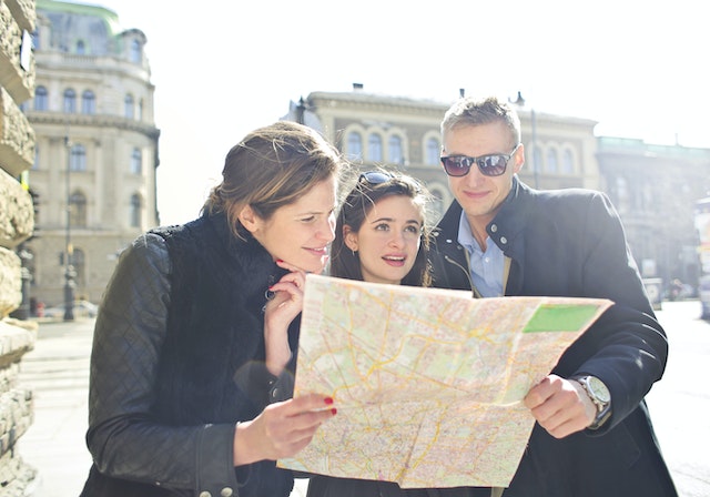Three people are looking at a map together on a sunny day in an urban environment with historical buildings in the background.