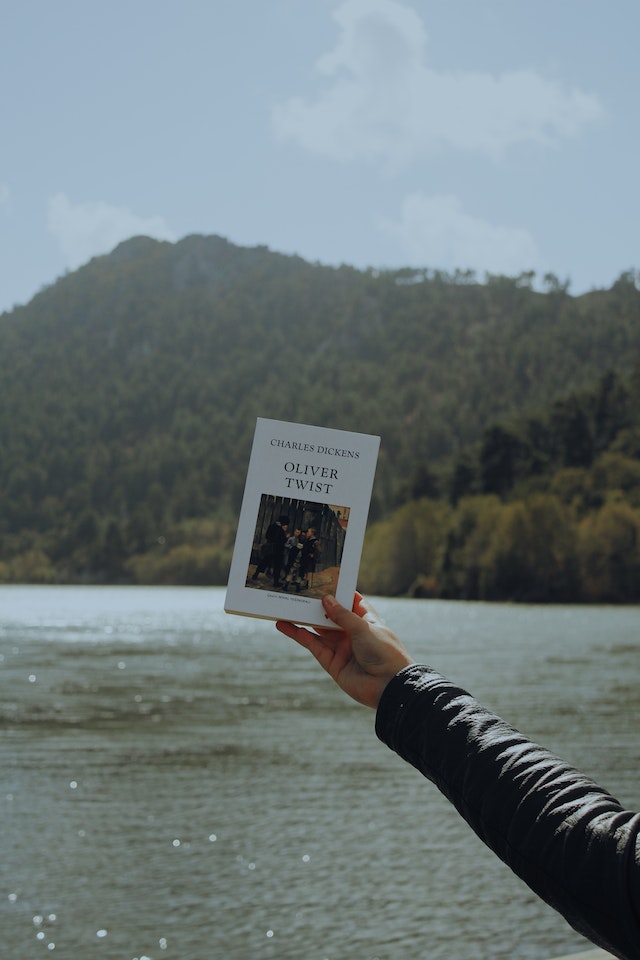 A person holds up "Oliver Twist" by Charles Dickens in front of a scenic backdrop with water and a forested mountain under a cloudy sky.