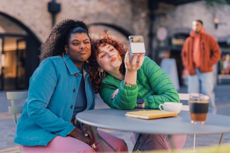 Two individuals are sitting at an outdoor cafe table, smiling and taking a selfie. Coffee, a notebook, and a passerby are also visible.